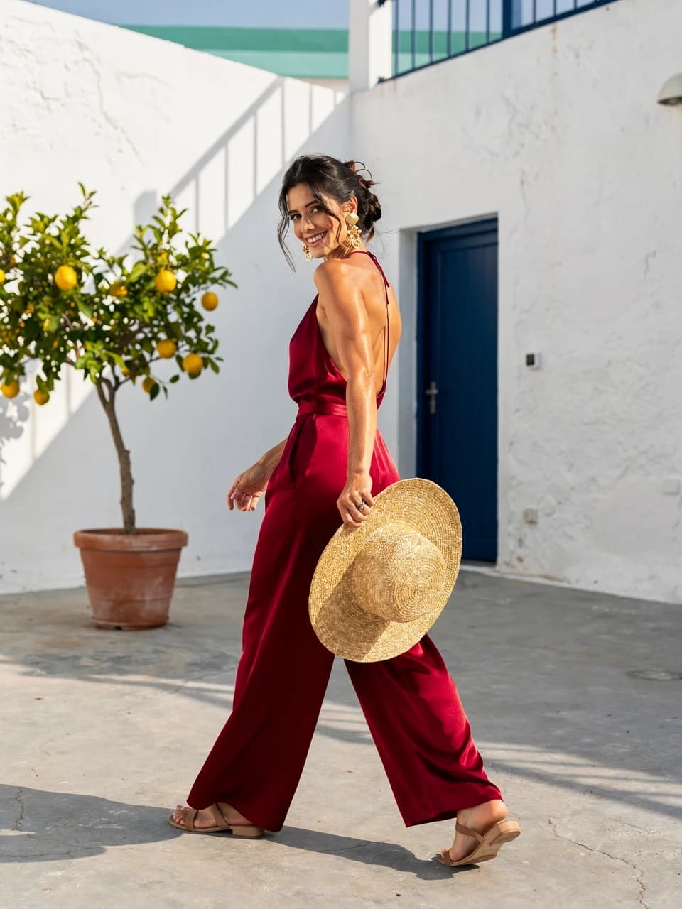 Woman in a scarlet silk jumpsuit turning mid-stride in a sunlit white courtyard.